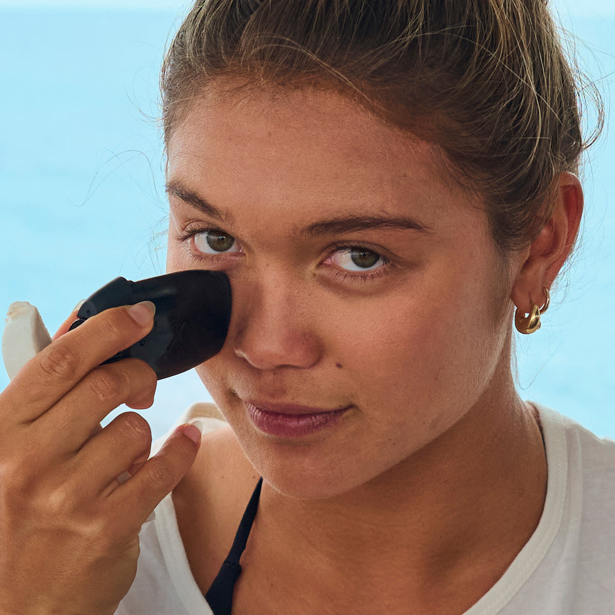 Woman applying sunscreen to her face with a black applicator against a clear blue sky.